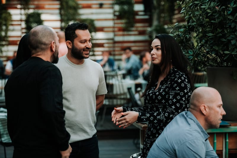 Four guests talking beside potted plants, one seated at a table, with diners behind them