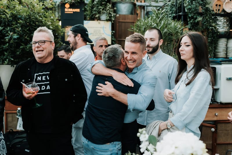 Two men hugging and laughing among guests in front of potted plants and a Sobranie London sign