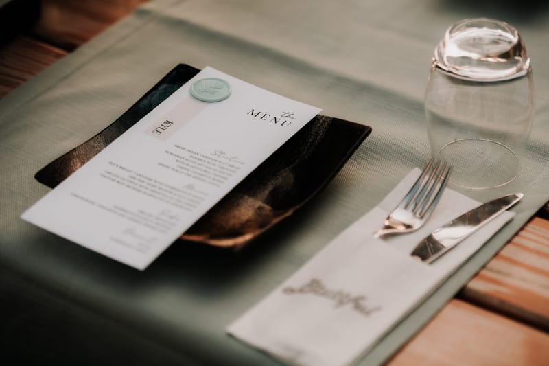 Named menu card on a dark plate beside an upturned water glass and a folded napkin
