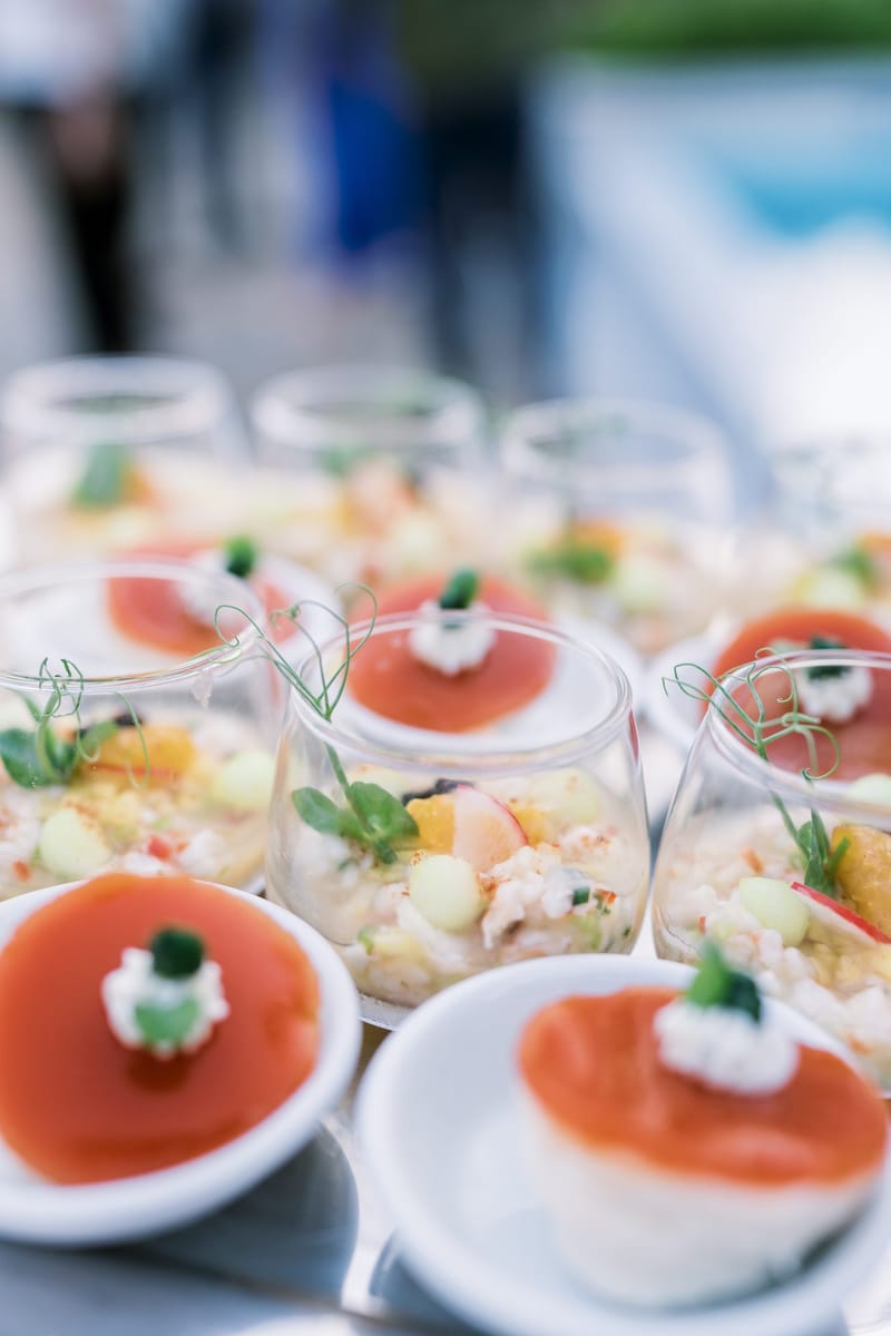 Rows of small canapés topped with red jelly and cream beside glasses of diced salad