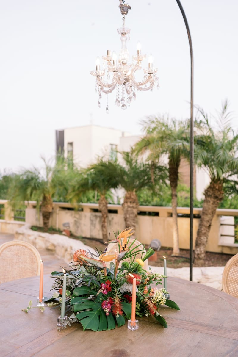Crystal chandelier hanging over a round wooden table of tropical leaves, flowers and tall candles among palms