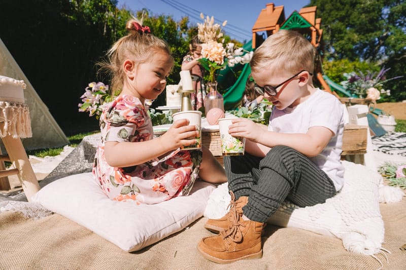 Two small children, one in a floral dress and one in glasses, holding cups on floor cushions at the picnic