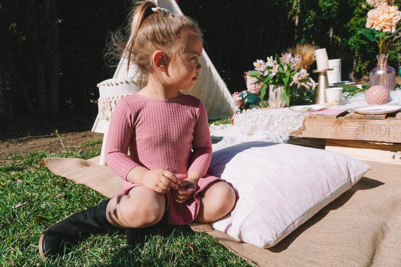 A girl in a pink dress sitting on a burlap rug beside a white floor cushion