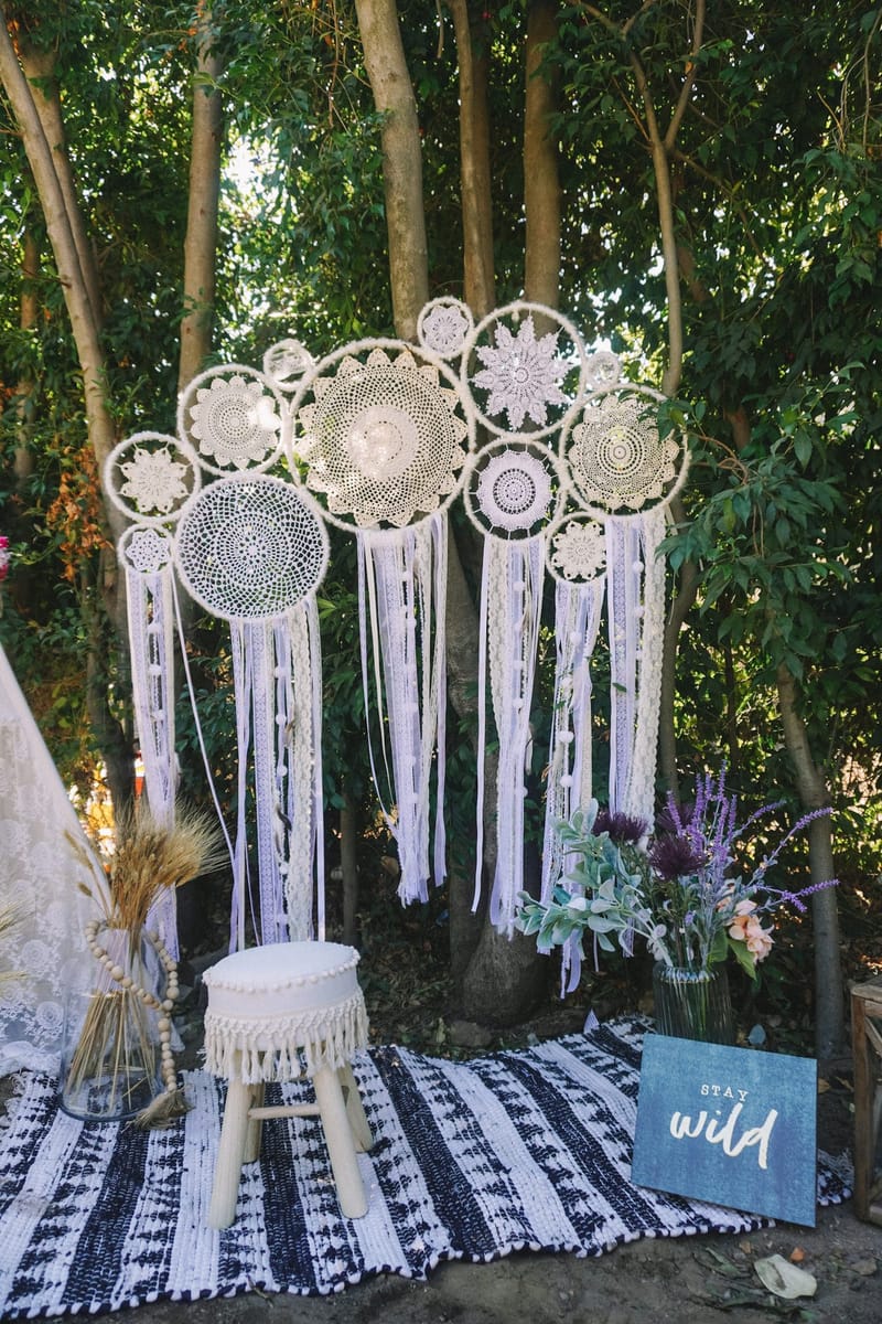 White macramé dreamcatchers hanging in the trees above a fringed stool, a striped rug and a STAY wild sign