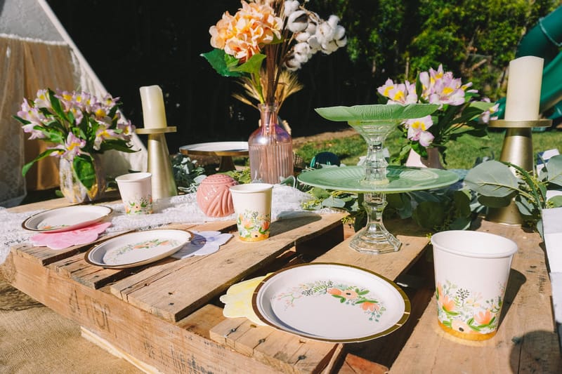 Pallet table set with floral plates, printed cups, a green glass cake stand and gold candle holders