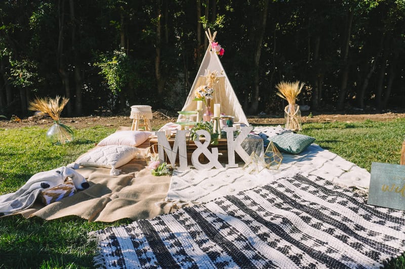 Picnic setup on the lawn with a white teepee, M&K letters, floor cushions and dried wheat in vases