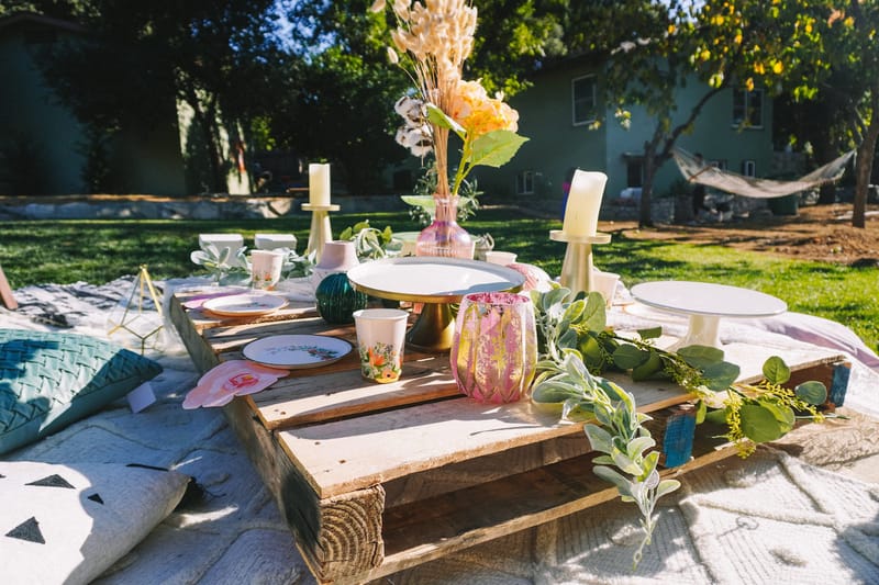 Low pallet table with cups, candles and a pink glass vase on a sunny lawn, a hammock in the background