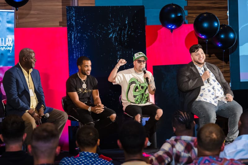 Four panelists seated on stage in front of red, blue and black panels with black balloons