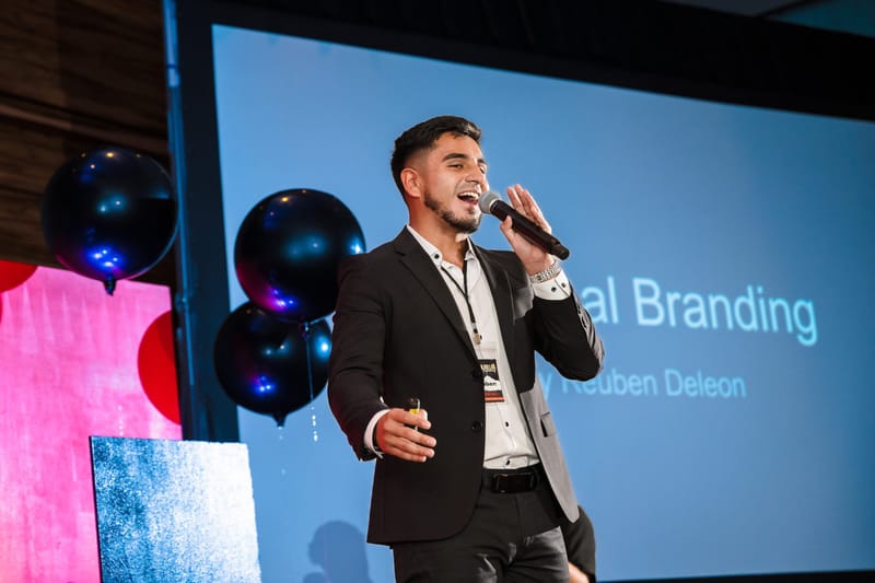 Speaker in a dark blazer with a microphone in front of a slide on branding