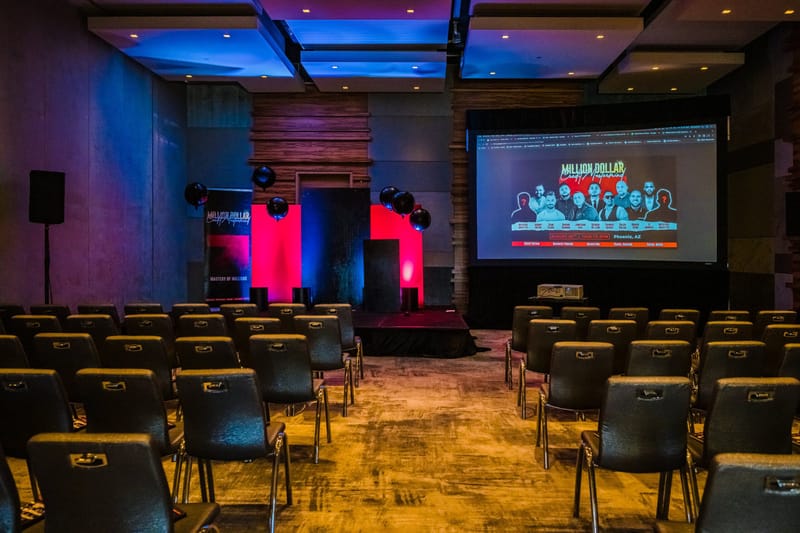 Empty rows of chairs facing a stage and a screen showing the speaker lineup