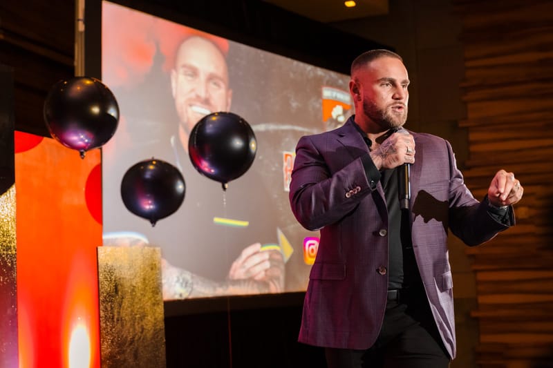 Speaker in a purple blazer gesturing on stage beside black balloons and a screen