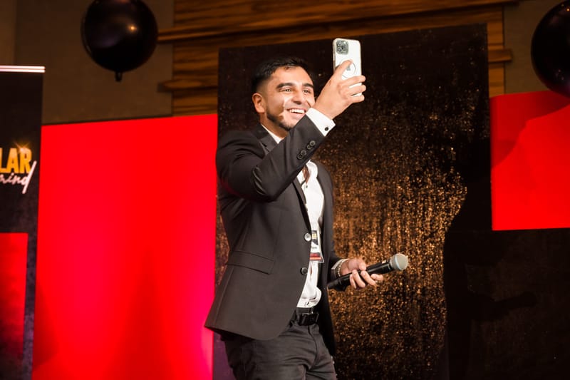 Speaker on stage taking a selfie with his phone in front of red and dark sequin panels