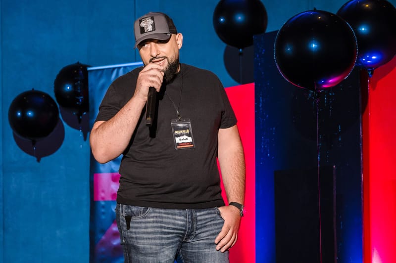 Man in a cap with a microphone and name badge in front of blue and red panels and black balloons