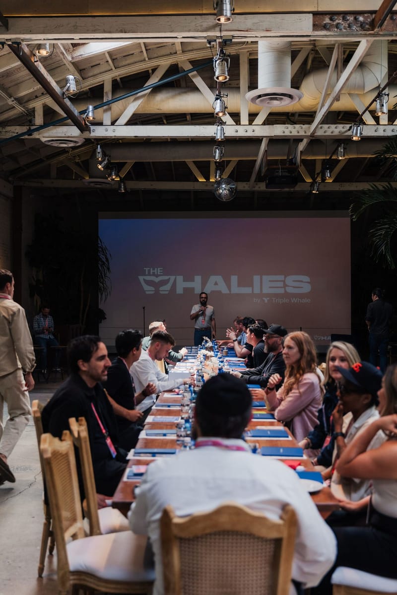 Long table of guests with notebooks and water bottles facing The Whalies screen under a warehouse roof