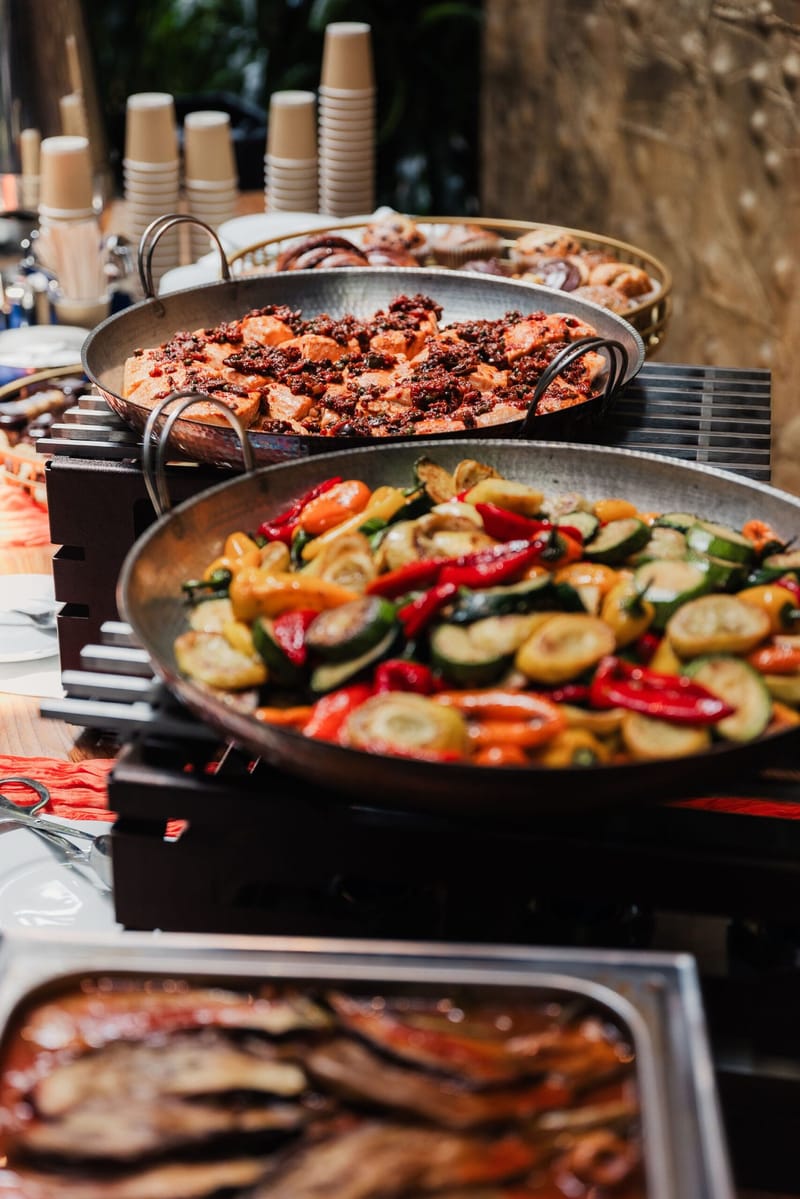 Large pans of roasted vegetables and salmon topped with tapenade on a buffet, with stacked paper cups
