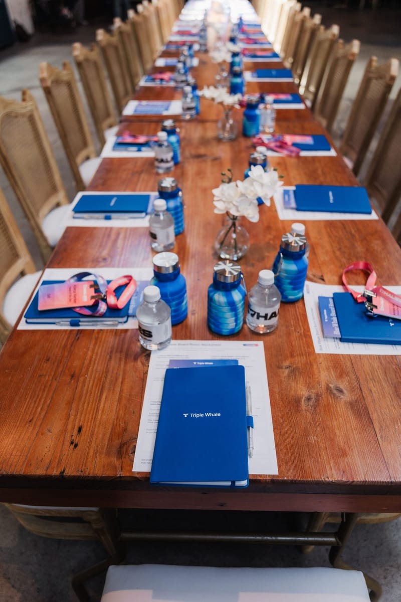 Long wooden table set with blue notebooks, blue water bottles, lanyards and white flowers