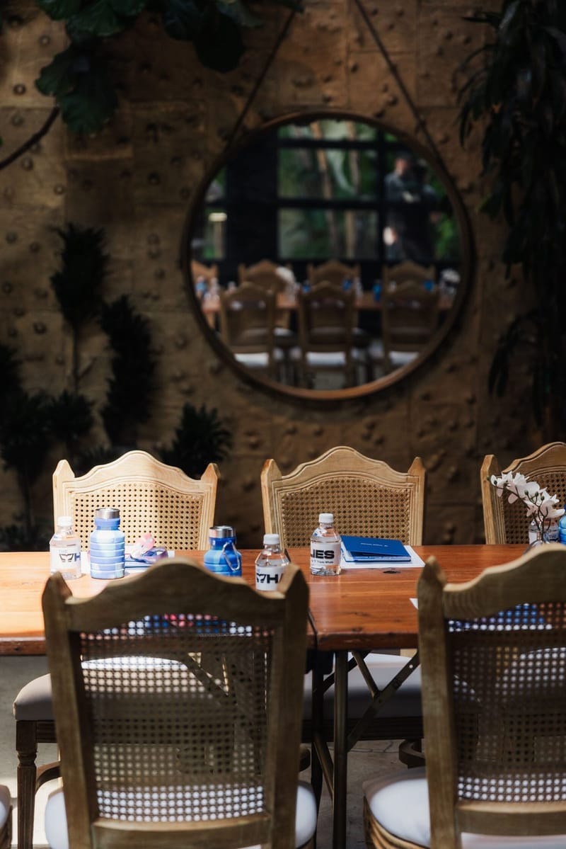 Rattan-back chairs and a table with water bottles in front of a round mirror on a stone wall