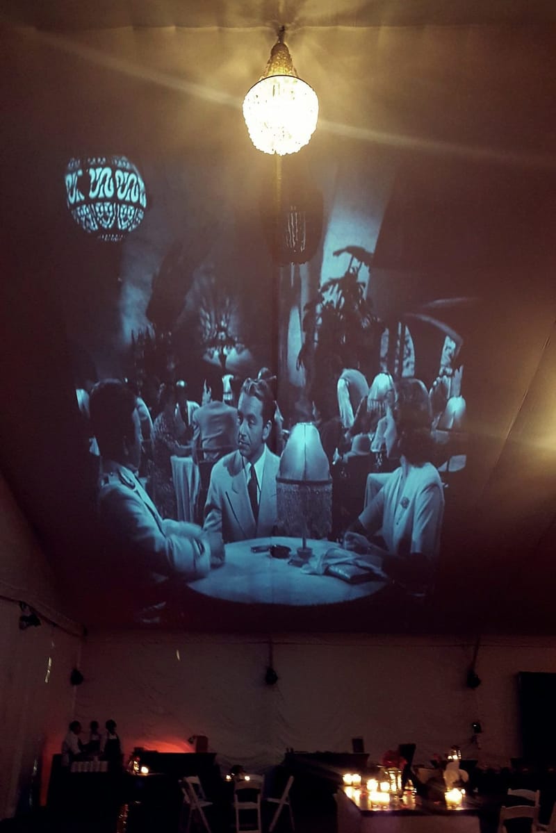 Black and white film scene projected across a white tent ceiling beside a lit chandelier, tables below