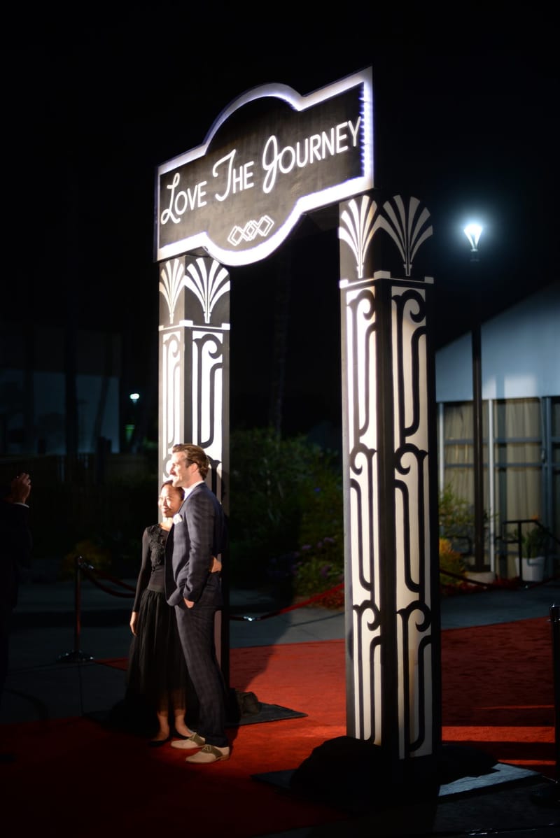 Two guests posing on a red carpet under a lit arched sign reading Love The Journey on art deco pillars