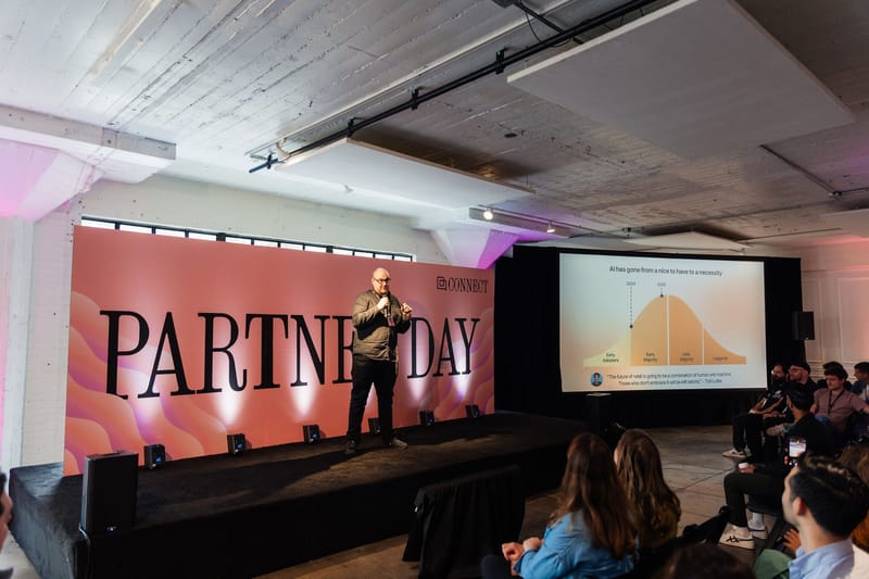 A speaker on stage in front of a large coral PARTNER DAY backdrop and a slide screen, under pink uplighting