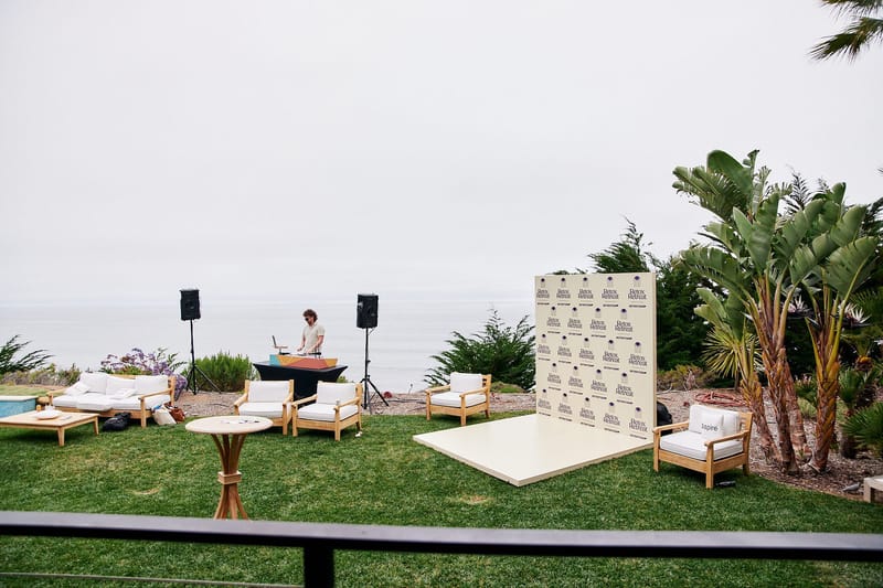 Lawn above the ocean with white lounge chairs, a DJ between speakers and a logo backdrop on a platform