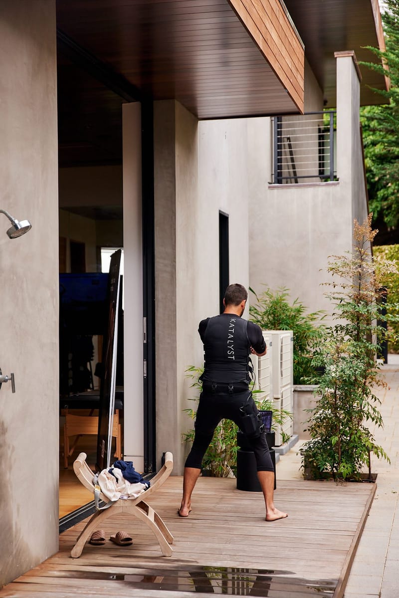 Barefoot man training in a black Katalyst suit on a wooden deck beside potted plants