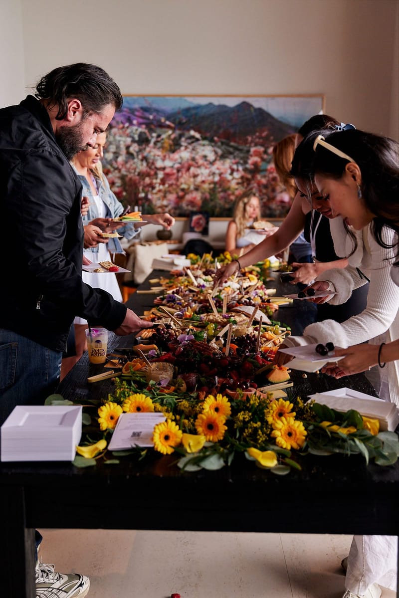 Guests serving themselves from a long grazing table edged with yellow flowers below a painting of a flowering hillside
