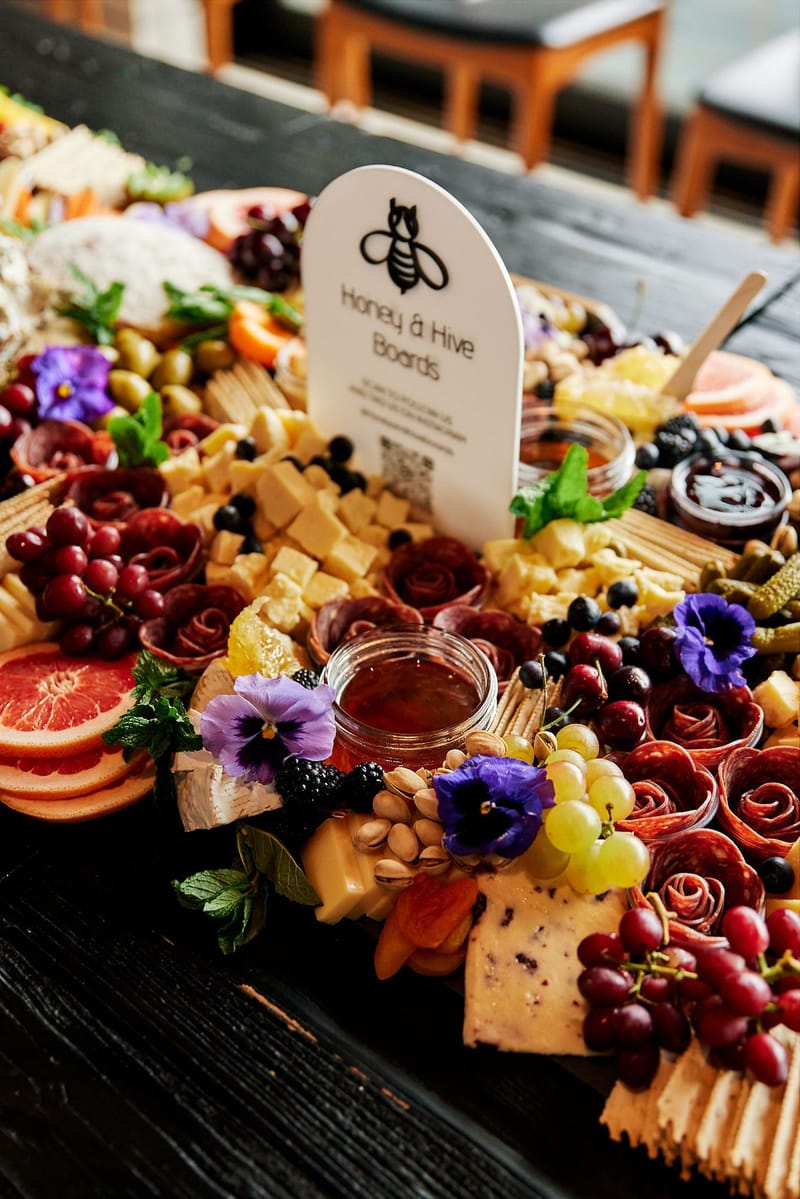 Honey & Hive Boards sign standing in a spread of cheese, salami roses, grapes and purple edible flowers
