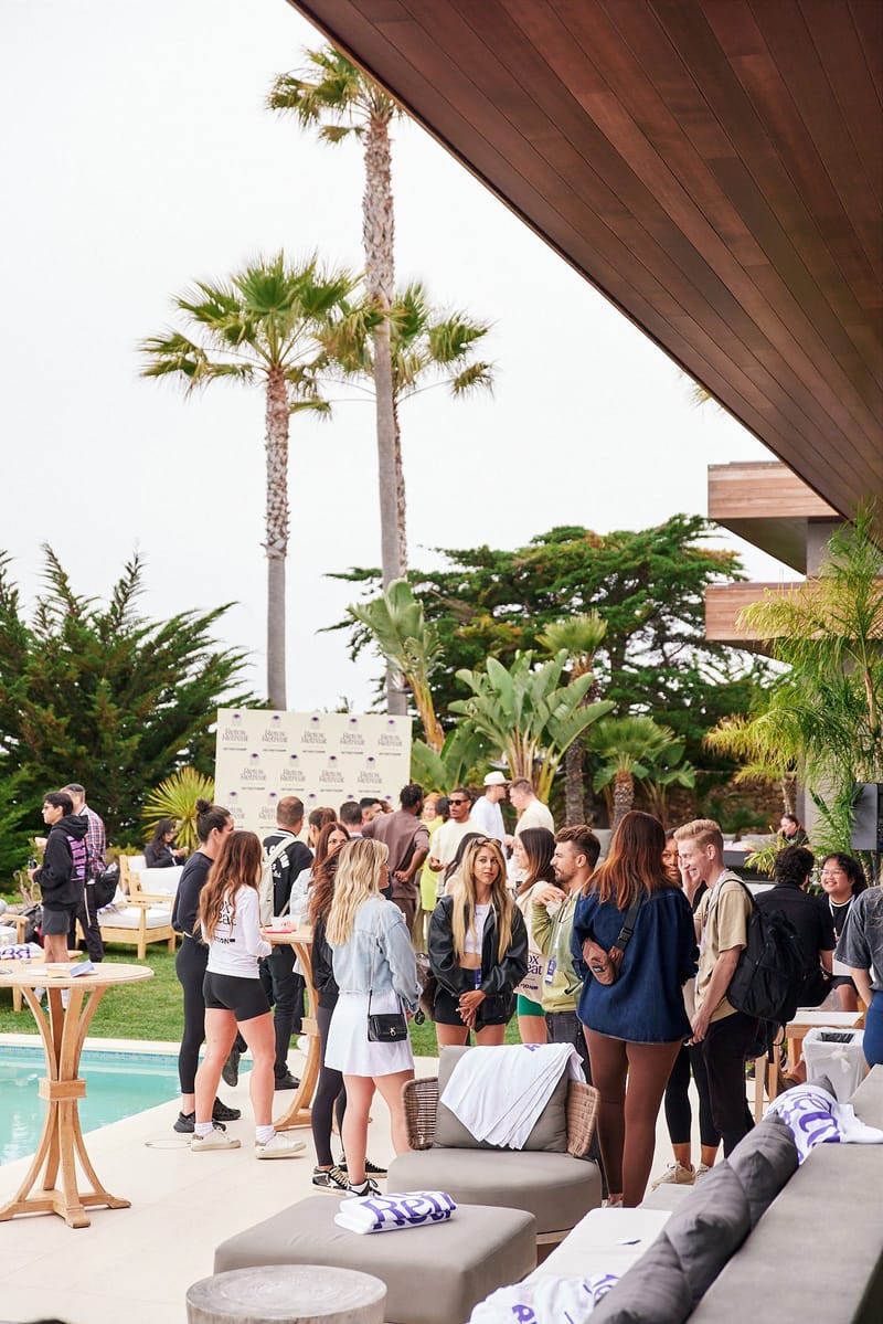 Guests mingling by the pool under palm trees, with white sofas and a logo backdrop behind them