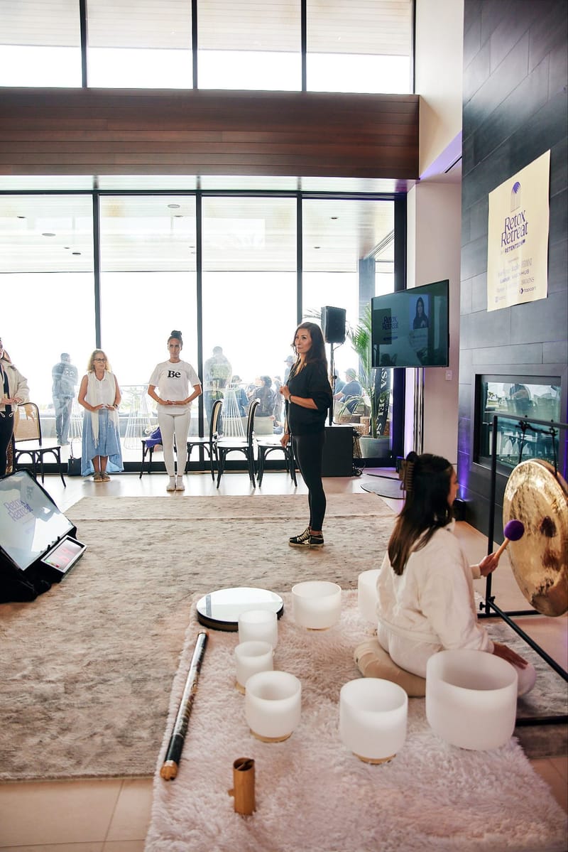 Practitioner playing a gong beside crystal singing bowls on a rug as guests stand and watch