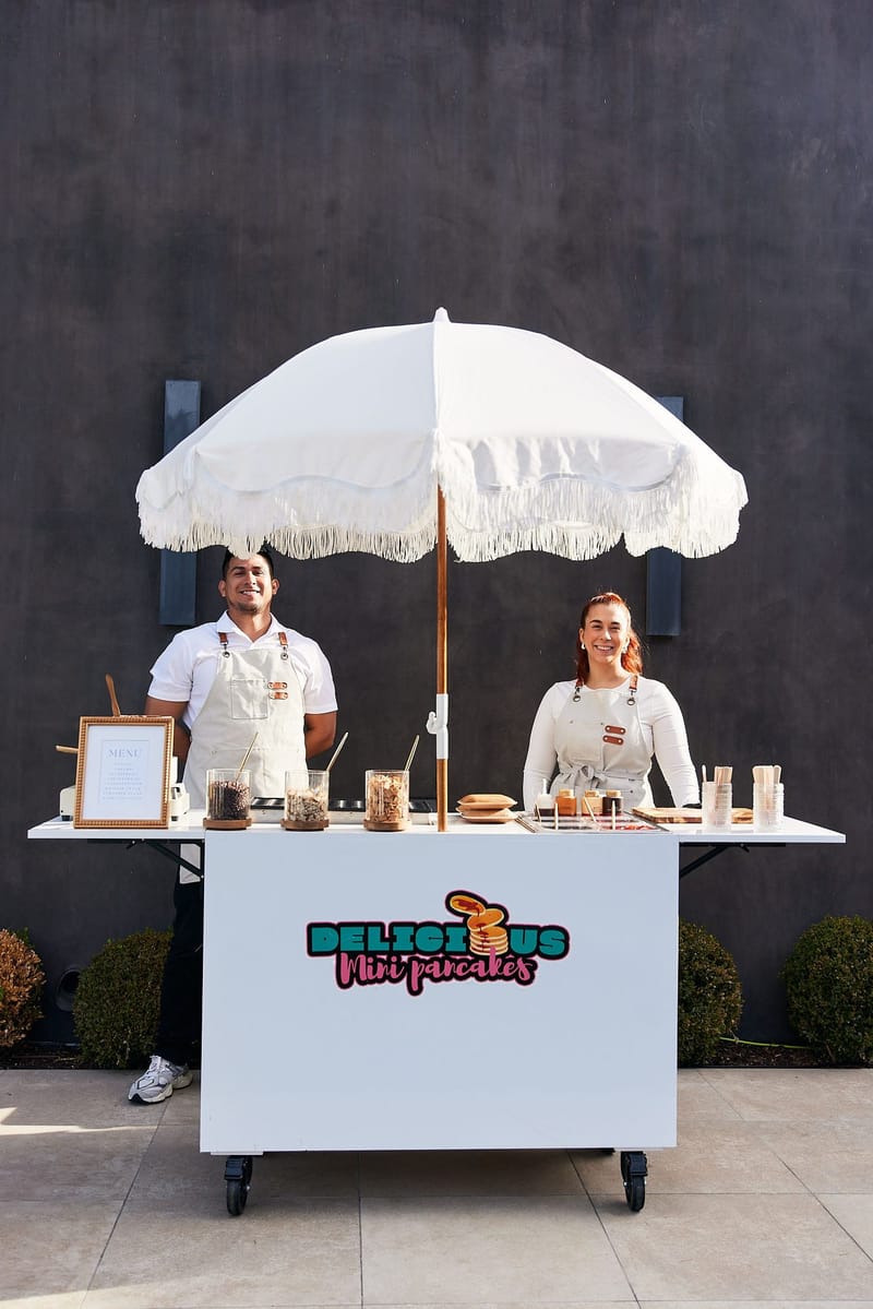 Two servers in aprons behind a mini pancakes cart with a fringed white umbrella