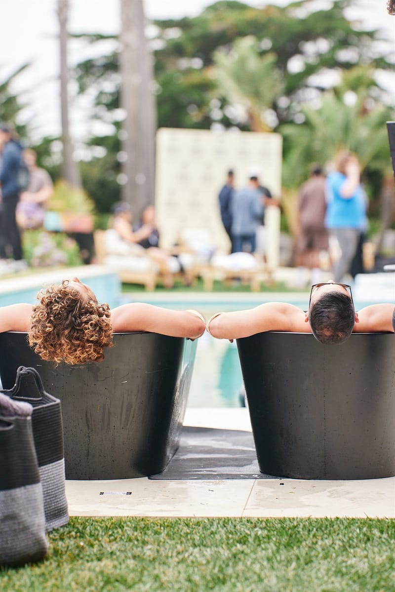 Two guests sitting in black ice bath tubs facing the pool and the lawn