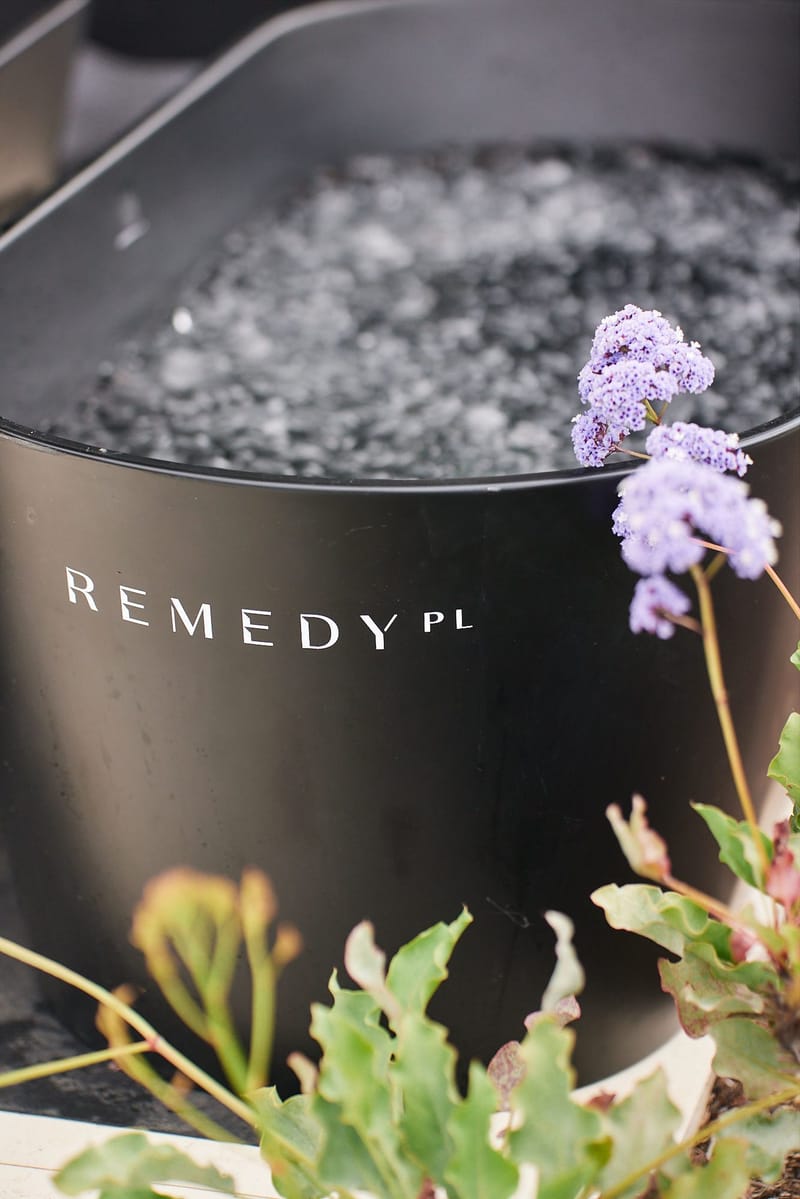 Black ice bath tub printed with REMEDY PL, filled with ice behind purple flowers