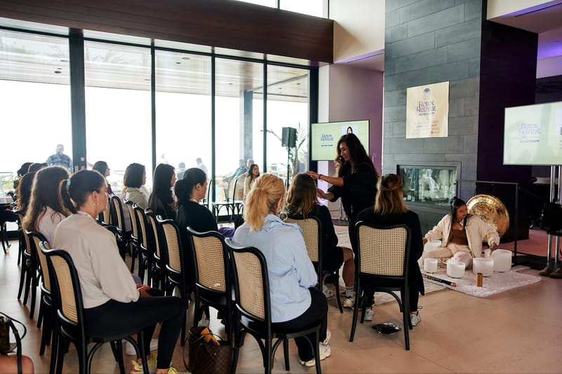 Rows of seated guests watching a speaker and a practitioner with a gong and bowls by a fireplace