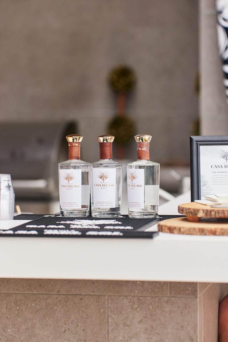 Three bottles of Casa Del Sol tequila blanco on a bar beside a framed Casa Del Sol card