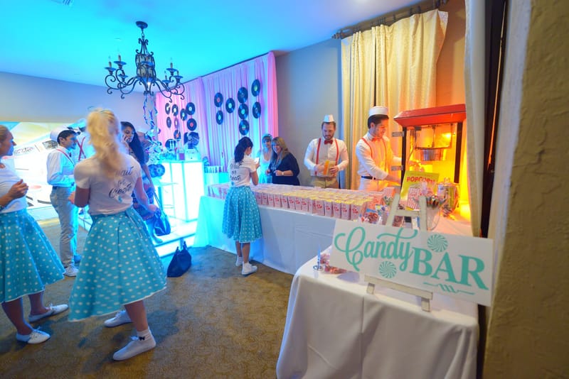 People in paper hats and red suspenders making popcorn at a long table behind a Candy Bar sign