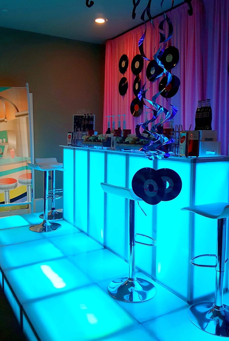 Blue glowing bar and floor with white stools, Coke bottles, vinyl records on pink drapes and a printed diner scene