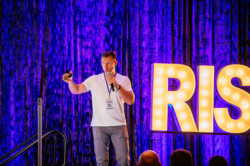 Speaker in a white T-shirt and lanyard holding a microphone beside lit RISE marquee letters