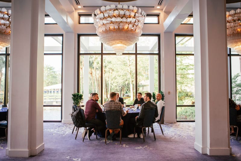 Guests seated at a round table under a crystal chandelier beside tall windows and white columns