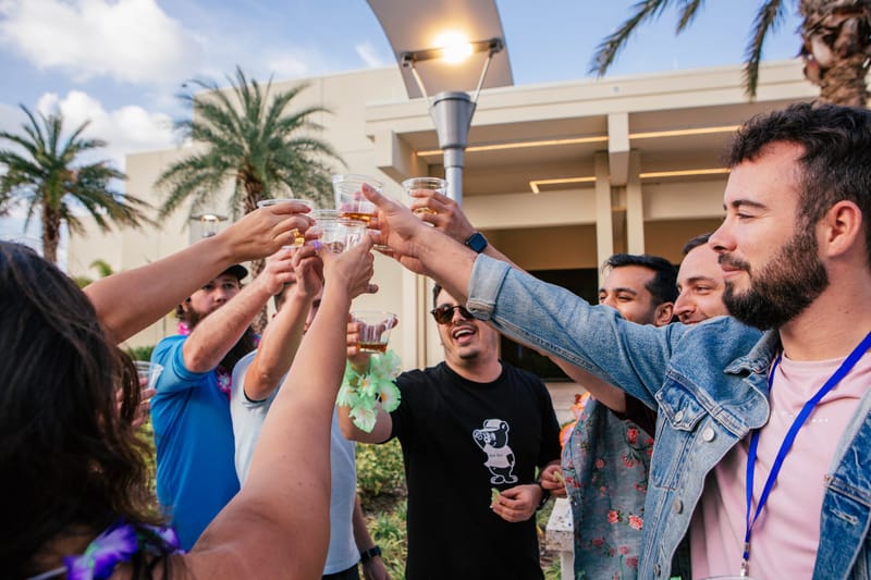 Guests wearing leis and lanyards raising plastic cups together in a toast beside palm trees