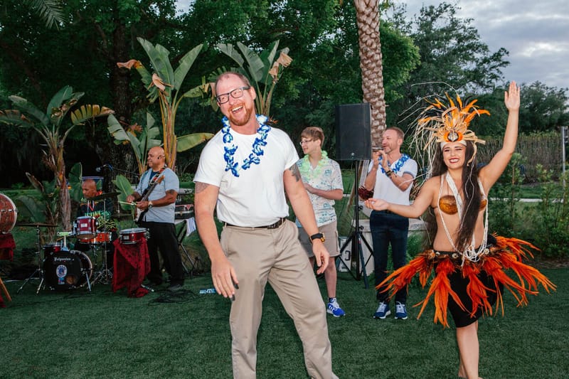 Guest in a blue lei dancing with a performer in a feathered headdress as a band plays behind