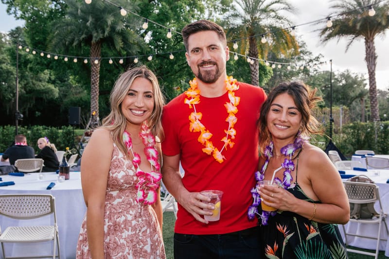 Three guests in flower leis holding drinks in front of string lights and round tables