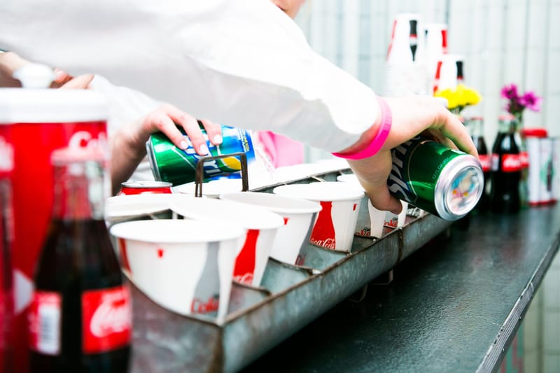 Two people pouring cans of Sprite into Coca-Cola paper cups set in a metal carrier