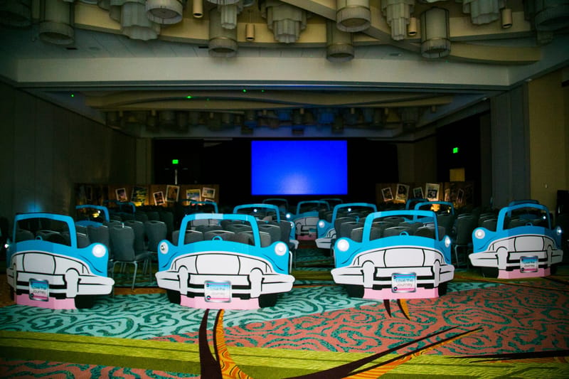 Blue classic car cutouts with Love the Journey plates in front of rows of chairs facing a large screen