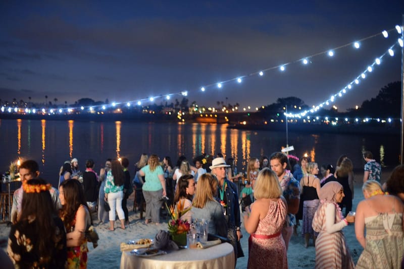 Guests talking on the sand at night under string lights reflected in the water
