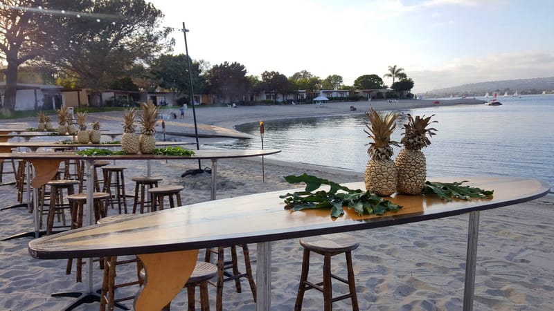 Surfboards used as high tables with wooden stools, pineapples and tropical leaves beside the water
