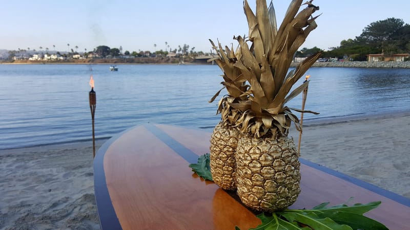 Two pineapples on tropical leaves on a wooden surfboard table, a tiki torch and the water behind