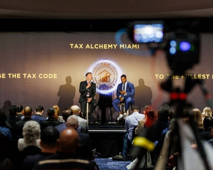 Two speakers on stage below Tax Alchemy Miami lettering, with a video camera in the foreground
