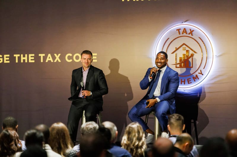 Two speakers seated on stools in front of a glowing round Tax Alchemy logo