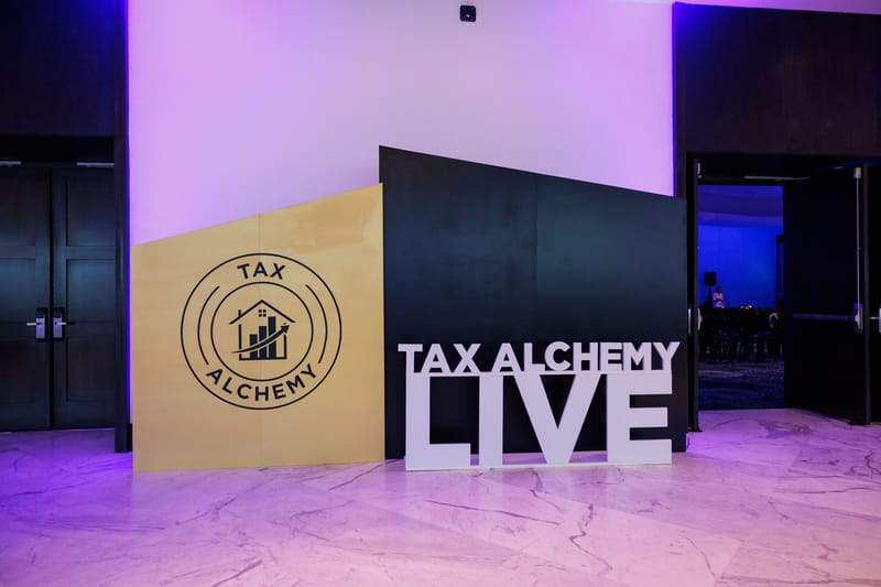 TAX ALCHEMY LIVE letters in front of a wood and black logo panel near doors lit in purple